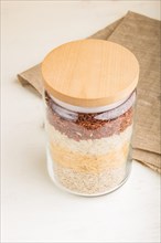 Glass jar with different kinds of rice poured in layers on white background. side view, close up.