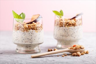 Yoghurt with plum, chia seeds and granola in a glass and wooden spoon on gray and pink background.