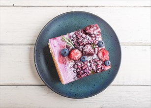 Berry cake with milk cream and blueberry jam on blue ceramic plate on a white wooden background.