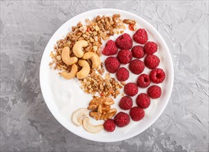 Yoghurt with raspberry, granola, cashew and walnut in white plate on gray concrete background and