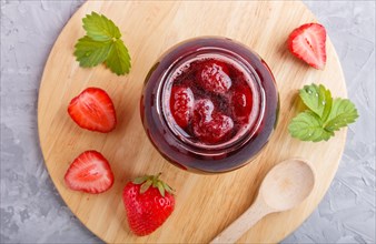Strawberry jam in a glass jar with berries and leaves on gray concrete background. Homemade, top