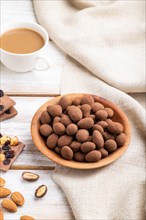 Almond in chocolate dragees in wooden plate and a cup of coffee on white wooden background and