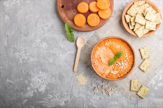 Carrot cream soup with sesame seeds and snacks in wooden bowl on a gray concrete background with