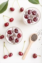 Yoghurt with cherries, chia seeds and granola in glass with wooden spoon on white wooden background