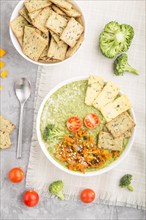 Green broccoli cream soup with crackers and cheese in white bowl on a gray concrete background and