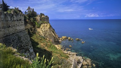 Steep cliffs and old fortress walls overlook the clear blue sea under a partly cloudy sky, sea