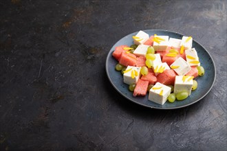 Vegetarian salad with watermelon, feta cheese, and grapes on blue ceramic plate on black concrete