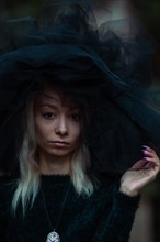 Mysterious white young blonde woman in a dark setting wear a large black tulle hat and veil and a