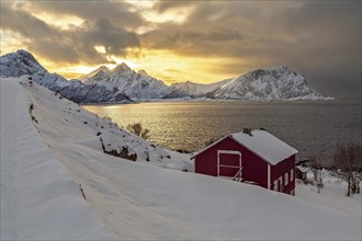 Ein rotes Haus vor einem winterlichen Sonnenuntergang mit verschneiten Bergen auf den Vesteralen