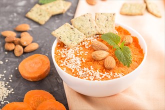 Carrot cream soup with sesame seeds, almonds and snacks in white bowl on a black concrete