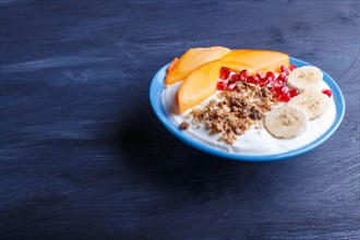 A blue plate with greek yogurt, granola, persimmon, banana, pomegranate black wooden background.