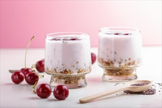 Yoghurt with cherries, chia seeds and granola in glass with wooden spoon on pink and white