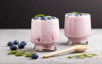 Yoghurt with blueberry and sesame in a glass and wooden spoon on gray and black background. side