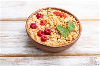 Wheat flakes porridge with milk, raspberry and currant in wooden bowl on white wooden background.