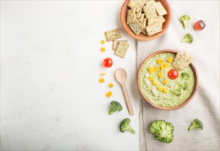 Green broccoli cream soup with crackers and cheese in wooden bowl on a white wooden background and