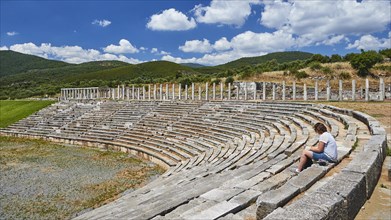 Person sitting in ancient stadium under clear blue sky with mountains in the background,