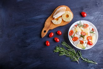 Chicken fillet salad with rosemary, pineapple and cherry tomatoes on dark blue wooden background.