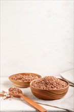 Two wooden bowls with unpolished brown rice and wooden spoon on a white wooden background and linen