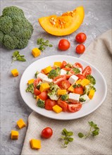 Vegetarian salad with broccoli, tomatoes, feta cheese, and pumpkin on white ceramic plate on a gray
