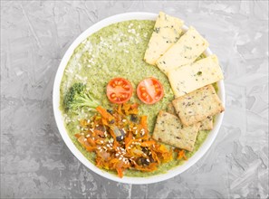 Green broccoli cream soup with crackers and cheese in white bowl on a gray concrete background. top