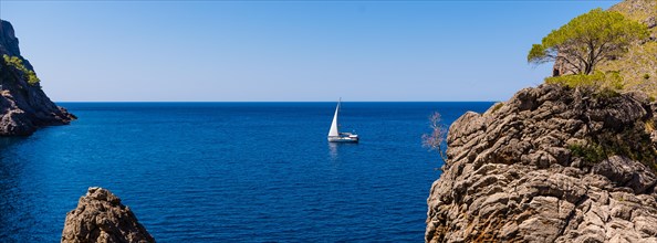 A sailing boat glides on calm blue waters between rocky shores under a clear sky, Majorca