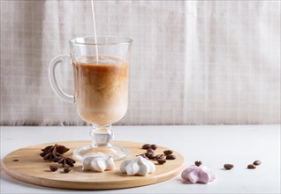 Glass cup of coffee with cream poured over and meringues on a wooden board on a white background.