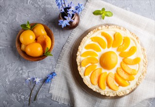 Round peach cheesecake and ceramic vase with blue flowers on a linen napkin on a gray concrete