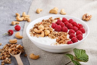 Yoghurt with raspberry, granola, cashew and walnut in white plate with wooden spoon on gray