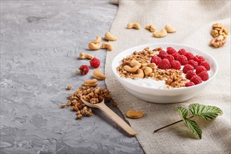 Yoghurt with raspberry, granola, cashew and walnut in white plate with wooden spoon on gray