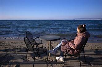 Young woman looking at the sea, lonely, alone, coat, table and chairs, beach bar, beach, Peraia,