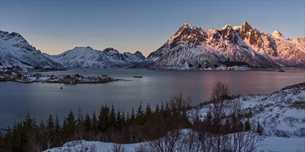 Die untergehende Sonne beleuchtet schneebedeckte Berge an einem ruhigen See, Lofoten