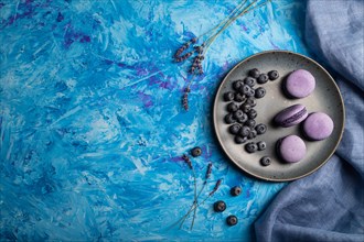 Purple macarons or macaroons cakes with blueberries on ceramic plate on a blue concrete background