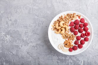 Yoghurt with raspberry, granola, cashew and walnut in white plate on gray concrete background and