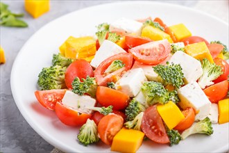 Vegetarian salad with broccoli, tomatoes, feta cheese, and pumpkin on white ceramic plate on a gray