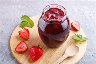 Strawberry jam in a glass jar with berries and leaves on gray concrete background. Homemade, close