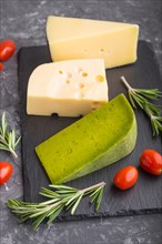 Green basil cheese and various types of cheese with rosemary and tomatoes on black slate board on a