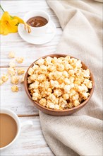 Popcorn with caramel in wooden bowl and a cup of coffee on a white wooden background and linen