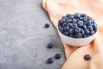 Fresh blueberry in white bowl and orange pastel textile on gray concrete background. side view,