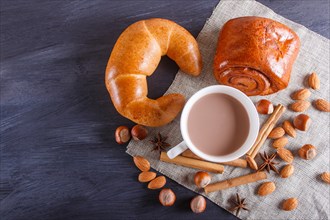 A cup of hot chocolate with nuts, buns and spices on linen napkin and black wooden background. copy