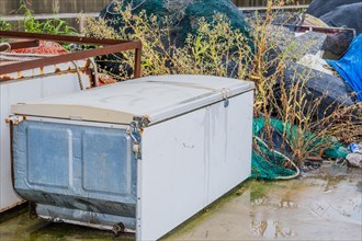 Old abandoned refrigerator laying on wet concrete in front of weeds and other debris on rainy