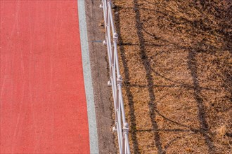 Looking down onto red bike path next to metal guardrail casting its shadow on brown grassy area in