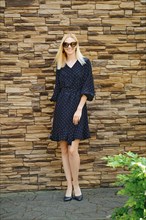 Happy fashionable woman in dotted loose dress posing in front of brick wall