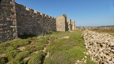 Historical ruins of an ancient wall under a clear blue sky with surrounding vegetation, sea