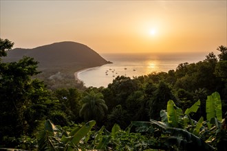View from a mountain onto a secluded bay with a sandy beach and mangrove forest. The sun sets over