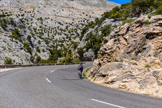 A cyclist rides along a winding mountain road surrounded by imposing rock faces under a clear blue