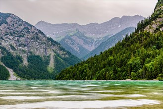 A picturesque mountain lake with green water surrounded by wooded mountains under a cloudy sky,
