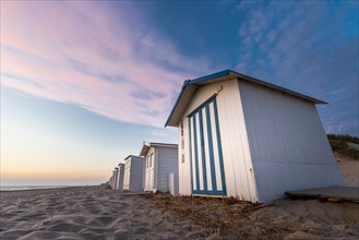 Evening mood with a view of white beach huts with blue stripes by the sea under a pink cloudy sky,
