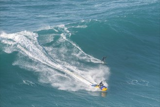A surfer rides a high wave and is accompanied by a jet ski, Nazare, Portugal, Europe