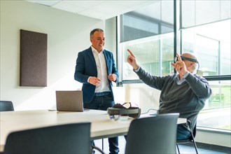Aged manager sitting and using VR goggles next to a employee in a office