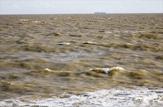 Brown choppy sea carrying sediment in suspension with distant container ship, North Sea, Suffolk,
